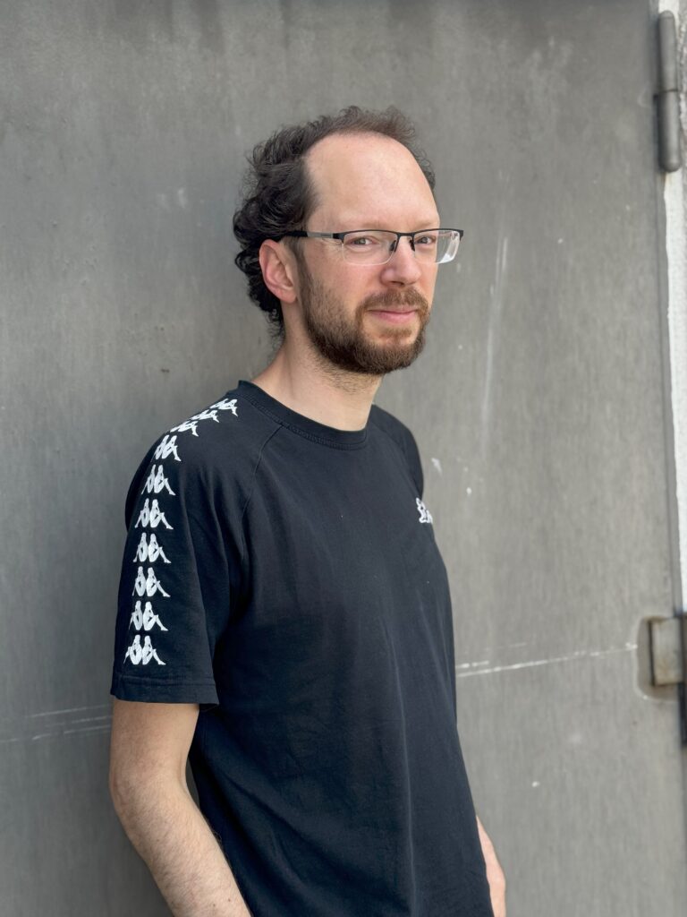 Portraitfoto von Christian: Er hat kurze, dunkelbraune Haare und einen kurzen Vollbart. Er trägt ein schwarzes T-Shirt.