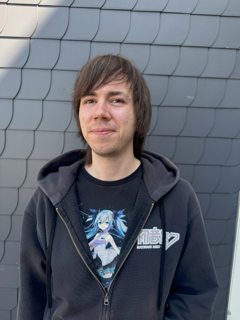 Portraitfoto von Max: Er hat schulterlange, dunkelbraune Haare. Unter einer Sweatshirtjacke trägt er ein schwarzes T-Shirt. Darauf befindet sich ein Bild des japanischen Pop-Idols "Hatsune Miku".