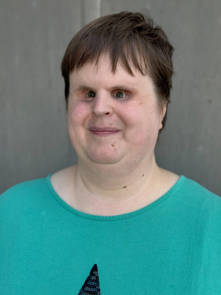 Portraitfoto von Nathalie: Sie steht vor einer grauen Wand und hat kurze, mittelbraune Haare. Auf ihrem grünen T-Shirt ist die obere Spitze eines schwarzen Sterns erkennbar.