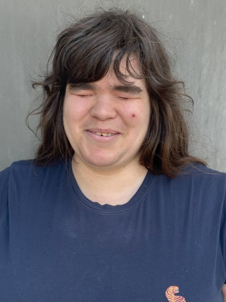 Portraitfoto von Tanja: Sie hat dunkelbraune Haare bis zur Taile und trägt ein dunkelblaues T-Shirt mit einem Tiger-Logo in orange.
