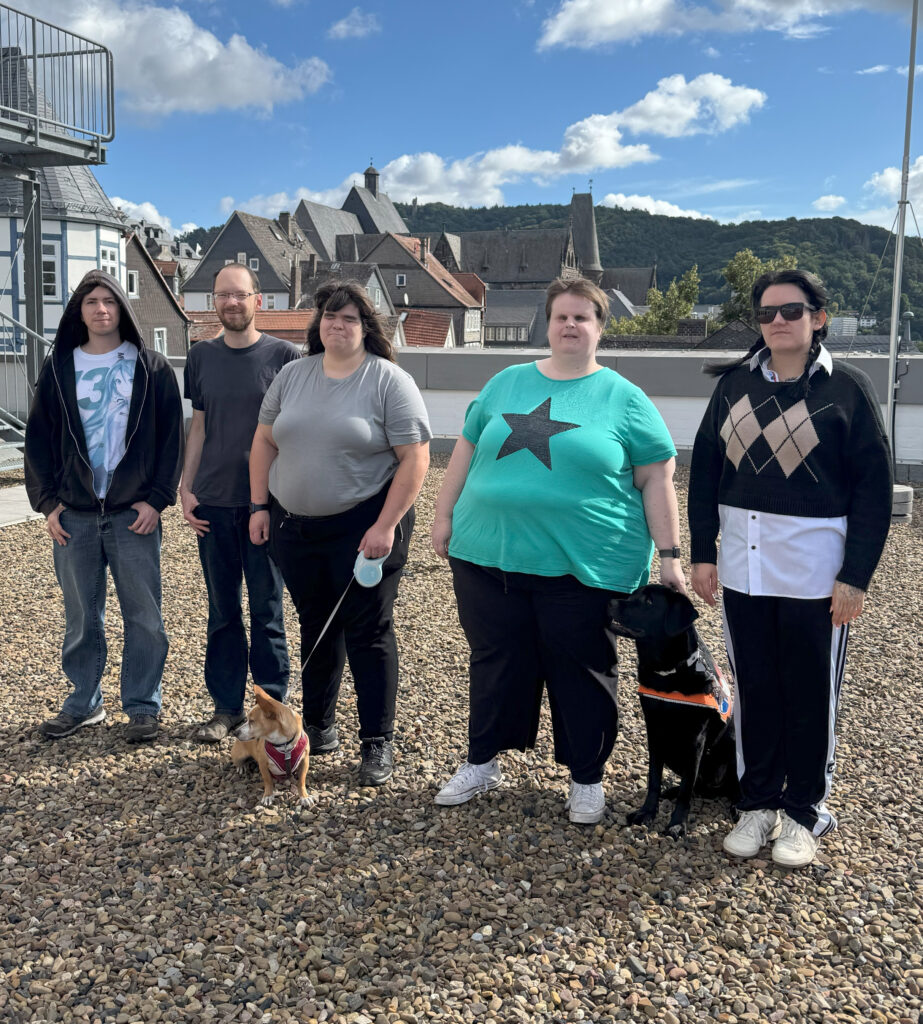 Gruppenfoto des inklusivio-Teams. Von links nach rechts: Max, Christian, Tanja, Nathalie, Angelina. Zu Füßen sitzen die Hunde Timon und Odin.